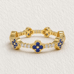 May include: A gold-colored ring featuring a repeating pattern of four-leaf clover designs with blue gemstones. The ring also has small white gemstones between the clover designs. The ring is set against a white background.