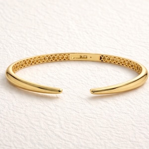 Puede incluir: Un brazalete de puño abierto de color dorado. El brazalete tiene un diseño liso y redondeado con un hueco abierto. La parte superior del brazalete presenta un patrón calado decorativo. El brazalete se muestra sobre una superficie blanca.