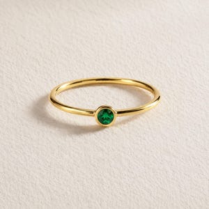Puede incluir: Un anillo dorado con una pequeña gema redonda de color verde esmeralda en el centro. La banda del anillo es delgada y pulida, reflejando la luz. El anillo se muestra sobre una superficie blanca.