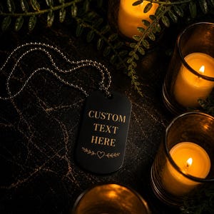 May include: A black dog tag necklace with the text "CUSTOM TEXT HERE" in gold lettering, accented with a heart and leaf design. The tag is on a silver ball chain, surrounded by lit candles in glass holders and greenery.