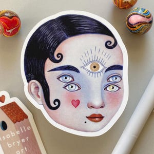 May include: A sticker depicting a face with multiple eyes and a third eye on the forehead. The face has dark hair, rosy cheeks, and a small heart. The sticker is next to a house-shaped sticker and colourful marbles.