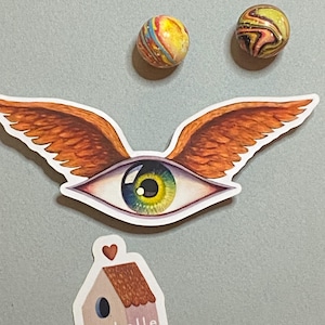 Op de afbeelding: Een sticker met een groot oog met oranje vleugels en een groene iris. De sticker bevindt zich op een grijze achtergrond met twee kleurrijke knikkers. Onder de sticker bevindt zich een kleine sticker van een huis met de tekst "isabelle bryer art" erop.