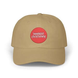 Peut inclure: Casquette de baseball beige avec une visière incurvée. La casquette présente un écusson circulaire rouge brodé des mots "Imminent Countdown!" en police blanc cassé. La casquette est en tissu doux.