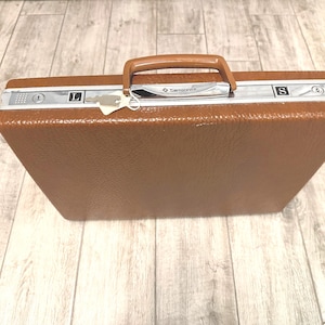 May include: A brown Samsonite briefcase with a brown handle and silver accents. The briefcase has a key attached to the lock and the letters "L" and "S" are visible on the silver trim.