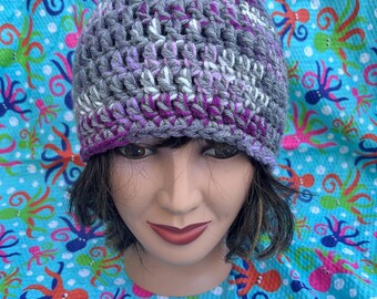 May include: A crocheted hat in shades of gray, white, and purple. The hat has a slouchy fit and is made with a textured stitch.