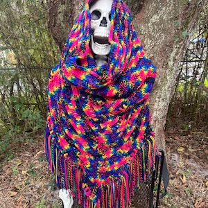 May include: A colorful, crocheted shawl with a vibrant mix of pink, blue, yellow, and green yarn. The shawl features a fringed edge and is draped over a white skull, creating a unique and eye-catching display. The shawl is a triangular shape.