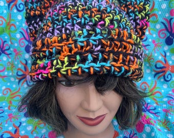 May include: A colorful crocheted beanie with cat ears. The beanie is made with a variety of bright colors, including pink, green, blue, and orange. The beanie is black with a multi-colored yarn.