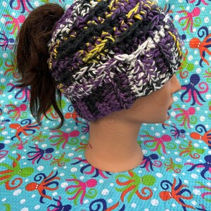 Può includere: Un berretto lavorato a maglia di colore nero, bianco, viola e giallo con un foro nella parte superiore per una coda di cavallo.