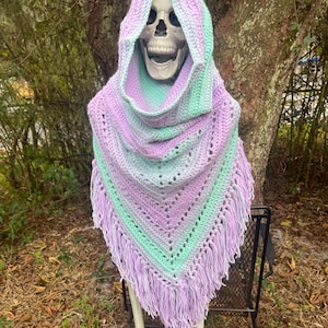 May include: A crocheted hooded shawl in shades of lavender, mint green, and gray, with a fringed edge. The shawl is draped over a skeleton, showcasing the garment's design and texture. The hood is up, and the shawl is triangular.