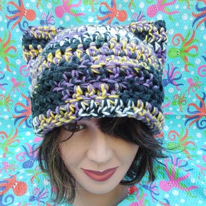 May include: A handmade crochet hat with cat ears. The hat features a mix of colors, including purple, yellow, white, and dark blue. The hat is shown on a mannequin head. The background is a light blue fabric with a colorful octopus pattern.