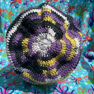 May include: A handmade crochet hat in a non-binary pride color scheme. The hat features a spiral design with black, white, yellow, and purple yarn. The hat is displayed on a blue fabric with a repeating octopus pattern.