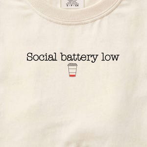 May include: Ivory-colored crew neck t-shirt with the text "Social battery low" and a small coffee cup graphic. The shirt has a Comfort Colors tag and a simple, minimalist design.
