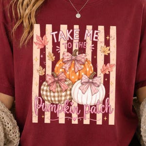 Peut inclure: Un t-shirt à manches courtes bordeaux avec un motif de trois citrouilles ornées de nœuds, entourées de feuilles d'automne et de rayures verticales. Le texte "TAKE ME TO THE Pumpkin patch" est écrit dans un mélange de polices avec des étoiles.
