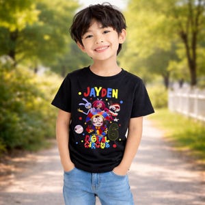 May include: A black t-shirt featuring the name "JAYDEN" in colorful letters above a graphic of cartoon characters from "The Amazing Digital Circus". The number "6" is also displayed. The child is wearing blue jeans.
