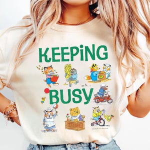 May include: A cream-colored t-shirt featuring the green text KEEPING BUSY and a collection of colorful illustrations depicting various anthropomorphic animals engaged in different activities, including cats, a bear, and a fox in clothing.