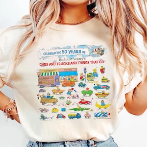 May include: A cream-colored t-shirt featuring a colorful graphic print celebrating 50 years of Richard Scarry's Cars and Trucks and Things That Go. The design includes a toy store storefront and various whimsical vehicles driven by animal characters.