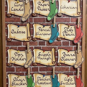 May include: A classroom job chart featuring a brick-patterned board with twelve scroll-shaped paper cutouts labeled with roles like Line Leader, Librarian, and Caboose. Each scroll is paired with a colorful paper sock cutout bearing a name.