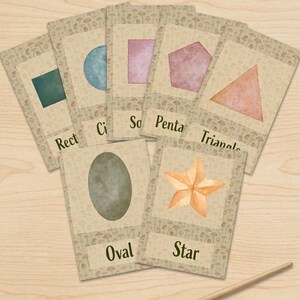 May include: A set of seven educational flashcards featuring watercolor shapes including a rectangle, circle, square, pentagon, triangle, oval, and star, each labeled with its name. A wooden paintbrush lies below the cards on a light wood surface.