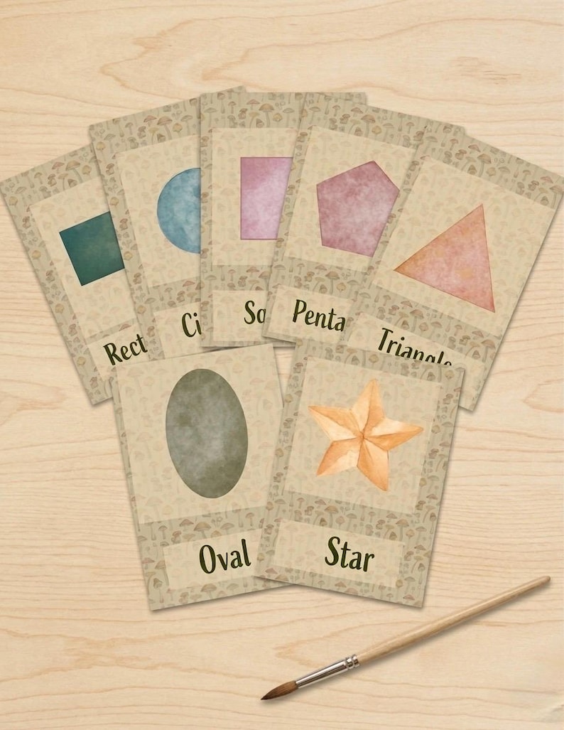May include: A set of seven educational flashcards featuring watercolor shapes including a rectangle, circle, square, pentagon, triangle, oval, and star, each labeled with its name. A wooden paintbrush lies below the cards on a light wood surface.