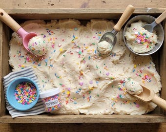 Può includere: Un vassoio di legno riempito con impasto color bianco sporco ricoperto di zuccherini colorati. Sono inclusi un porzionatore con manico rosa, un porzionatore in metallo con manico in legno, un cucchiaio di legno, una ciotola blu con zuccherini, una ciotola in metallo e un barattolo con l'etichetta TOPPINGS.