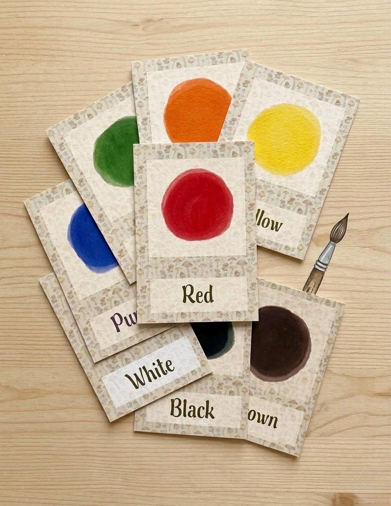 May include: A collection of color cards with watercolor circles in various hues, including red, yellow, and blue. Each card has the color name printed on it. A paintbrush is also visible.