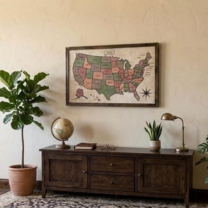 May include: A framed map of the United States with muted green and red states hanging above a dark wood media console. The console features two cabinets and two central drawers, topped with a globe, a book, a small plant, and a brass lamp.