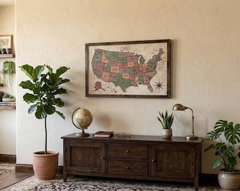 May include: A framed map of the United States with muted green and red states hanging above a dark wood media console. The console features two cabinets and two central drawers, topped with a globe, a book, a small plant, and a brass lamp.