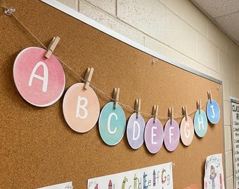 Può includere: Una ghirlanda orizzontale composta da dieci ritagli di carta circolari, ognuno dei quali mostra una lettera maiuscola bianca dalla A alla J. I cerchi sono di colore pastello e appesi a uno spago sottile con piccole mollette di legno.