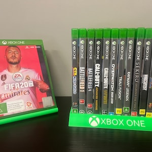 Könnte beinhalten: Eine Sammlung von Xbox One-Videospielhüllen, darunter "FIFA 20 Emirates", auf einem grünen Ständer. Die Hüllen zeigen verschiedene Spieletitel und Cover-Art. Der Ständer hat das Xbox One-Logo.