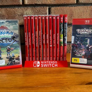May include: A collection of Nintendo Switch video game cases, including "Pokémon Legends: Arceus" and "Pokémon Legends: Nintendo Switch 2 Edition." The cases are red and feature game titles and artwork. A red stand with the Nintendo Switch logo holds the game cartridges.
