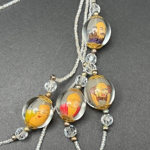May include: A multi-strand necklace featuring clear glass beads and larger spherical beads, each containing a hand-painted illustration of a distinct character with unique facial expressions and clothing, accented by gold-toned metal caps.