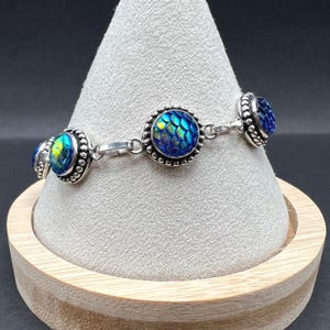 Può includere: Bracciale argentato con cabochon blu e verdi iridescenti con motivo a scaglie e bordo perlato. Il bracciale è esposto su un supporto a forma di cono di colore chiaro. I cabochon hanno un diametro di circa 1,5 cm.