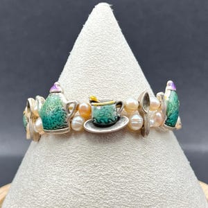 Könnte beinhalten: Ein Armband mit einer Reihe kleiner, runder, cremefarbener Perlen, die mit silberfarbenen Anhängern durchsetzt sind. Die Anhänger zeigen grün und violett emaillierte Teekannen, eine Teetasse auf einer Untertasse und Löffel in einem Muster.
