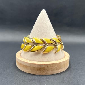 Puede incluir: Un brazalete vintage con diseño de hojas. El brazalete presenta hojas de esmalte amarillo brillante con detalles metálicos dorados. El brazalete se exhibe sobre un soporte en forma de cono beige, sobre un fondo oscuro. Una joya elegante.