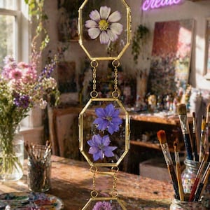 Op de afbeelding: Een verticale hangende decoratie met drie verbonden zeshoekige koperen frames. Elk frame bevat geperste bloemen tussen twee heldere glasplaten. Het bovenste frame bevat één witte bloem, het middelste twee paarse bloemen en het onderste drie kleine paarse bloemetjes.