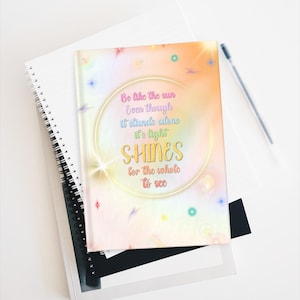 Puede incluir: Un cuaderno de espiral con una cubierta colorida con el texto "Be like the sun...SHINES for the whole to see". El diseño incluye un círculo dorado y colores pastel. Un bolígrafo descansa sobre el cuaderno.