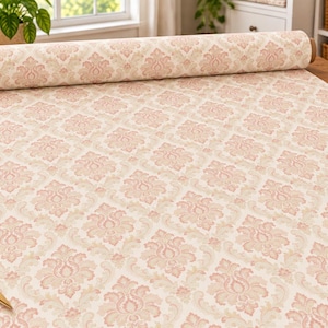 May include: A roll of fabric featuring a repeating damask pattern in soft pink and cream tones. The fabric is laid out flat, showcasing the intricate floral-inspired design. Nearby are gold-toned scissors, a white measuring tape, and a pin cushion.
