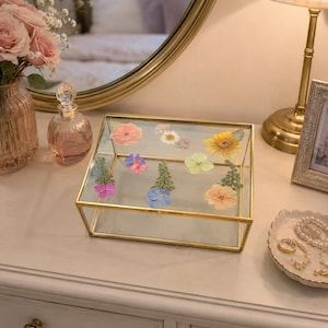 Könnte beinhalten: Eine rechteckige Glas-Aufbewahrungsbox mit einem dünnen goldfarbenen Metallrahmen. Der Deckel ist mit einer Auswahl an gepressten, getrockneten Blumen in verschiedenen Farben wie Rosa, Gelb, Blau und Grün auf der transparenten Fläche verziert.