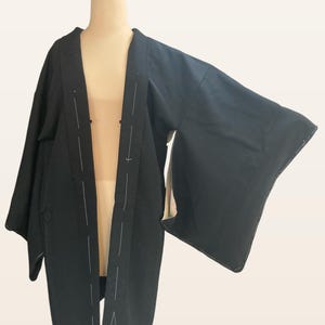May include: A black kimono-style robe with wide sleeves, displayed on a mannequin. The robe features white stitching along the edges and seams, with subtle geometric patterns near the hem and sleeves. The fabric appears to be a matte material.