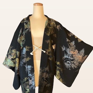 May include: A black haori jacket featuring a detailed landscape print with shades of green, blue, and gold. The garment has wide sleeves and is secured at the front with a white braided cord. The fabric displays a repeating pattern of trees.