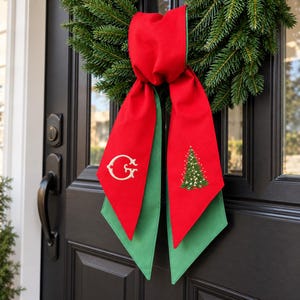 Peut inclure: Un nœud en tissu rouge et vert suspendu à une couronne. Le pan gauche présente une lettre G dorée brodée, tandis que le pan droit affiche un sapin de Noël vert brodé avec des détails blancs. Le nœud a de longues extrémités pointues.