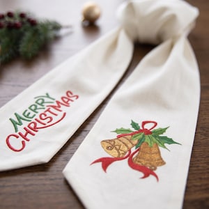 May include: An off-white fabric runner featuring embroidered text that reads Merry Christmas in green and red thread. The other end of the runner displays an embroidered design of two gold bells with green holly leaves and a red ribbon.