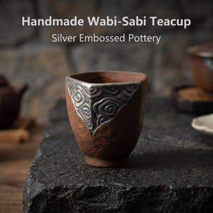 May include: A handmade Wabi-Sabi teacup made of silver embossed pottery. The teacup is brown with a silver embossed design on the rim. The text "Handmade Wabi-Sabi Teacup" and "Silver Embossed Pottery" are visible.