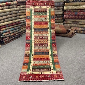 Op de afbeelding: Een lang, smal loperkleed met een levendig, veelkleurig geometrisch patroon met horizontale banden in rood, groen, blauw en crème. Het kleed toont ingewikkelde tribale motieven en bevat een label met de tekst 254x82 8251.