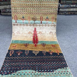 Op de afbeelding: Een lang, rechthoekig loperkleed met horizontale banen in diverse kleuren, waaronder diepblauw, olijfgroen, crème en turkoois. Het kleed is versierd met herhalende patronen van gestileerde bomen, kleine bloemmotieven en figuren van herten.