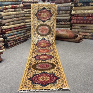 May include: A long rectangular runner rug featuring a cream-colored background with repeating geometric medallions in shades of red, blue, and dark brown. The rug displays the text 8269 354x79 in the upper right corner and has fringe ends.
