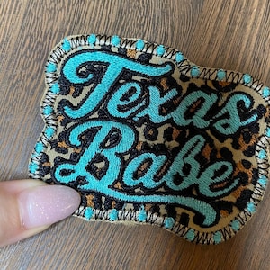 May include: Embroidered patch with the words "Texas Babe" in turquoise script. The patch has a leopard print background and a turquoise border with black stitching. The patch is rectangular with rounded corners.