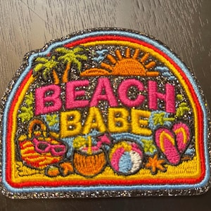 May include: A semi-circular embroidered patch with a glittery black background featuring the text BEACH BABE in pink and yellow. The design includes palm trees, an orange sun, a beach bag, a coconut drink, a beach ball, and pink flip-flops.