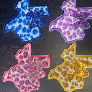 May include: Four Texas-shaped patches featuring leopard print patterns in blue, purple, pink, and gold. Each patch is accented with a matching glittery bow embellishment attached to the right side, finished with a stitched border around the edges.