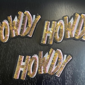 May include: Three embroidered patches featuring the word HOWDY in gold sequins and light pink thread. Each patch has a black border and a slanted, stylized font. The letters are filled with alternating vertical rows of gold sequins and pink stitching.